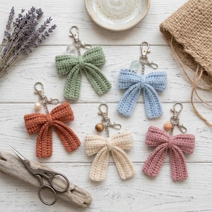 Peut inclure: Cinq porte-clés nœuds en crochet de couleurs vert sauge, bleu clair, terre cuite, crème et rose poudré. Chaque nœud est doté d'un fermoir mousqueton en métal argenté, d'une courte chaîne et d'une perle décorative fixée sur le dessus.