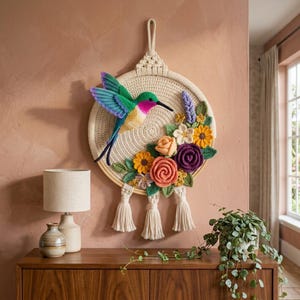 May include: A circular wall hanging featuring a crocheted hummingbird with green, purple, and yellow wings, surrounded by crocheted flowers in shades of orange, yellow, and purple, all mounted on a round frame with three hanging cream-colored tassels.