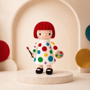 May include: A crocheted doll with bright red hair, wearing a white dress decorated with multicolored polka dots. The doll holds a small paintbrush in one hand and a miniature artist palette in the other, featuring black shoes and a light skin tone.