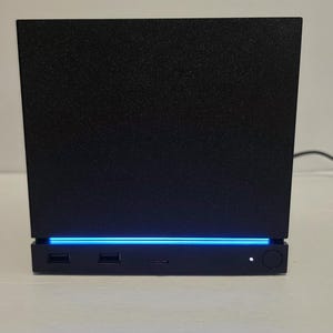 May include: A black rectangular electronic device with a textured finish. The front panel features a horizontal blue LED light strip, two USB ports, a central port, a small white indicator light, and a circular power button on the right side.