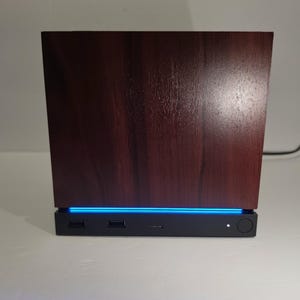 May include: A rectangular electronic device featuring a dark brown wood grain finish on the upper section and a black base. The base includes two USB ports, a small central port, a circular power button, and a bright blue LED light strip.