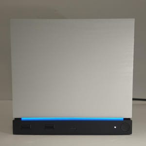 May include: A square, silver-colored electronic device featuring a smooth, flat front panel. The base is black and includes two USB ports, a small circular power button with a white indicator light, and a horizontal blue LED light strip.