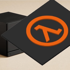 May include: A black cube-shaped computer case with a matte finish, featuring a small circular power button on the front panel. A flat, square black panel with an orange Greek letter lambda symbol inside an orange circle is positioned in front.