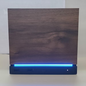 May include: A rectangular electronic device featuring a dark brown wood grain front panel. A horizontal strip of bright blue LED light glows beneath the wood panel. The black base contains two USB ports, a small slot, and a circular power button.