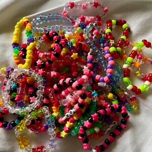 May include: A collection of colorful beaded bracelets in various designs. The bracelets feature a mix of star-shaped and round beads in shades of pink, yellow, green, blue, and black. Some bracelets have letter beads spelling out words.