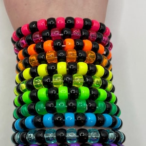 Può includere: Una pila di braccialetti di perline colorate in un arcobaleno di colori, tra cui rosa, arancione, giallo, verde, blu e viola. Ogni braccialetto è fatto di piccole perline rotonde e perline nere.