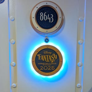 Könnte beinhalten: Eine weiße Tür mit zwei runden Plaketten. Die obere Plakette zeigt die Nummer 8643. Die untere Plakette lautet "Disney Fantasy Norwegian Fjords 2025". Die untere Plakette wird von blauem Licht beleuchtet.