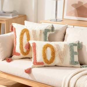 May include: Two off-white rectangular throw pillows with the word "LOVE" in colourful, textured letters. The pillows have tassels on the corners and are on a light-coloured sofa. The letters are in shades of yellow, pink, and green.