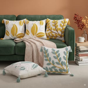 Puede incluir: Un conjunto de cojines decorativos con diseños bordados de hojas y flores en amarillo, azul y blanco. Los cojines están colocados sobre un sofá verde con una manta de punto crema. Cada cojín tiene borlas.