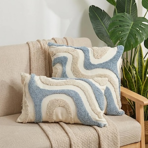 Könnte beinhalten: Zwei rechteckige Zierkissen mit einem wellenförmigen Muster in Hellblau und Beige auf cremefarbenem Grund. Sie liegen auf einem beigen Sofa mit einer passenden Decke. Die Kissen verleihen dem Raum Textur und Farbe.