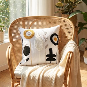 Puede incluir: Un cojín decorativo cuadrado blanco con un diseño floral en negro, amarillo y blanco, apoyado en una silla de mimbre. El cojín presenta formas de flores abstractas bordadas y está colocado sobre una manta de color crema.