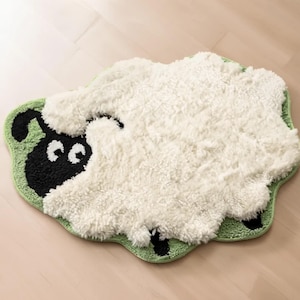 Puede incluir: Una alfombra de baño con forma de oveja con un cuerpo blanco y esponjoso y un borde verde. La cara de la oveja es negra con ojos blancos. La alfombra está diseñada para parecerse a una oveja de dibujos animados.