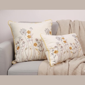 Könnte beinhalten: Zwei dekorative Zierkissen mit floralem Design. Die Kissen sind cremefarben mit gelbem Rand und weisen ein Muster aus gelben und grauen Wildblumen auf. Sie liegen auf einem grauen Sofa mit einer beigen Decke.