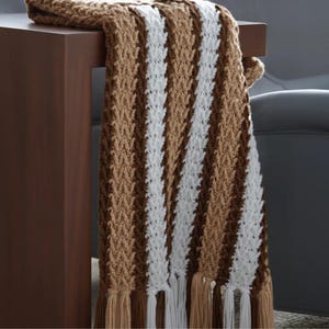 May include: A brown, white, and tan striped crochet blanket with fringe. The blanket is draped over a dark brown wooden table. The blanket's pattern is a series of vertical stripes. The fringe is tan and white.