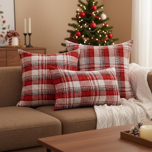 Könnte beinhalten: Drei rot-weiß karierte Zierkissen auf einem braunen Sofa. Die Kissen haben unterschiedliche Größen und Formen. Im Hintergrund steht ein Weihnachtsbaum mit roten Kugeln. Eine weiße Decke ist über das Sofa drapiert.
