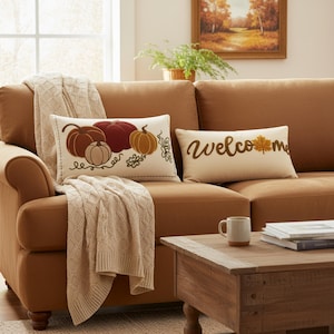 Può includere: Un divano marrone con due cuscini decorativi e una coperta beige. Un cuscino presenta zucche ricamate nei toni del marrone e dell'arancione. L'altro cuscino dice "Welcome" con una foglia. Un tavolino di legno contiene una tazza e dei libri.
