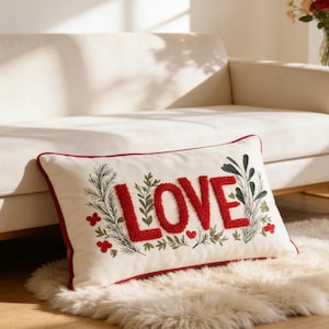 May include: Cream rectangular cushion embroidered with the word "LOVE" in red, textured letters. The cushion features green and red floral embroidery and a red border. It rests on a white faux fur rug, near a cream-colored sofa.