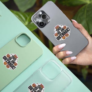 May include: A sticker with the text "THIS IS AN OUTRAGE!" and cat illustrations is affixed to a phone. The sticker is white with black text and orange cat faces. Two phone cases, one green and one clear, also display the sticker.