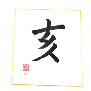 May include: Square artwork featuring a black calligraphy character on a white background, framed by a thin gold border. A small red seal is in the lower left corner. The artwork is displayed against a white backdrop.