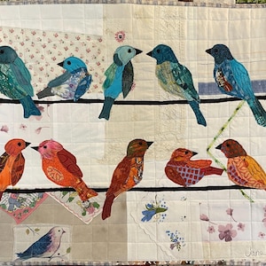 May include: A colourful quilt featuring a row of nine birds perched on a wire. The birds are made of fabric scraps and are in shades of blue, orange, and red. The quilt is made of various fabric scraps and has a white background. The quilt is signed "Jane H".