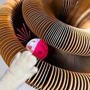 Op de afbeelding: Een witte kattenpoot reikt naar een roze en witte bal in een bruin, harmonica-achtig kattenspeeltje. Het speelgoed heeft een cirkelvormig ontwerp met meerdere lagen, waardoor een tunnelachtige structuur ontstaat. De bal heeft gaten en is ontworpen om mee te spelen.