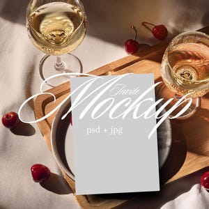 May include: A mockup scene featuring two wine glasses filled with a golden liquid, a plate, and a wooden tray. Red cherries are scattered around the scene. A light gray card with the text "Invite Mockup" is in the center.
