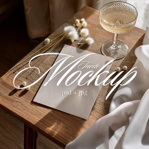 Könnte beinhalten: Eine Holzoberfläche mit einem Champagnerglas, einem Einladungs-Mockup und dekorativen Elementen. Das Einladungs-Mockup trägt den Text "Invite Mockup" und "psd + jpg". Die Szene umfasst Perlen und weißen Stoff.