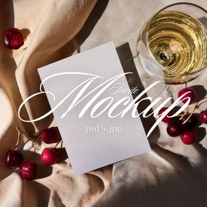 May include: A mockup scene featuring a blank white card with the word "Invite Mockup" in elegant script. The card is set on a beige fabric, accompanied by a glass of wine and fresh cherries. The image includes the text "psd + jpg".