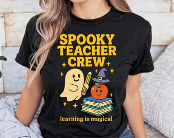 Op de afbeelding: Zwart T-shirt met een Halloween-thema. Het ontwerp toont een spook, potlood, appel met een heksenhoed en boeken, met de tekst "Spooky Teacher Crew" en "learning is magical".