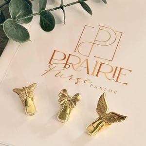 May include: Three gold-colored purse clips with butterfly, bow, and bird wing designs. The clips are arranged on a cream-colored surface with the text "PRAIRIE Purse Parlor" in gold.