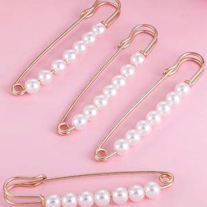 May include: Four gold-toned safety pins with a row of white pearls. The pins are arranged on a pink background. The pins are a decorative accessory.