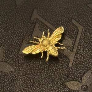 May include: A gold-colored bee-shaped pin. The pin has detailed wings, legs, and body segments. The pin is set against a dark, textured background with a subtle pattern.