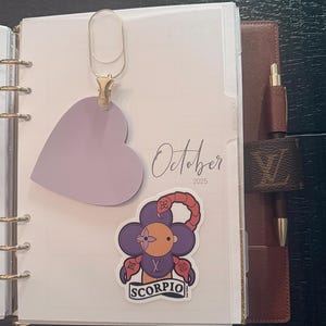 May include: A planner open to the October 2025 page, featuring a heart-shaped paper clip and a Scorpio sticker. The planner has a brown leather cover with a pen holder. The sticker is a purple flower with a face and the word "Scorpio".