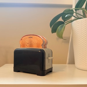 May include: A black and grey plastic toaster-shaped memo holder with two slices of toast-shaped paper sticking out of the top. The front slice of toast has the word notes printed in black lowercase letters on its textured, light brown surface.