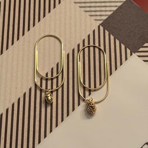 Può includere: Un paio di orecchini color oro. Ogni orecchino presenta un grande anello allungato con un piccolo ciondolo dettagliato appeso in basso. Un ciondolo è una piccola ghianda, l'altro è una pigna. Gli orecchini sono posti su uno sfondo a motivi.