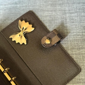 May include: A brown leather planner with gold-colored hardware and a gold-colored bow-shaped accent. The planner has a snap closure with a gold button. The interior features gold-colored rings for holding pages.