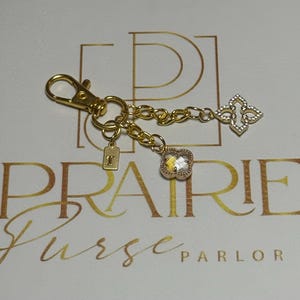 Puede incluir: Llavero dorado con mosquetón, cadena y tres colgantes. Los colgantes incluyen una pequeña etiqueta con la letra "T", un colgante cuadrado transparente con borde dorado y un colgante de trébol de cuatro hojas con piedras transparentes. El texto "PRAIRIE Purse PARLOR" es visible.