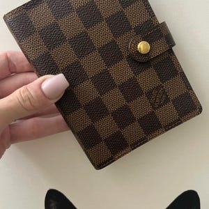 May include: A rectangular wallet featuring a brown and dark brown chequered pattern. The wallet includes a small tab closure secured with a circular gold-toned metal snap button. The exterior material has a textured finish throughout the surface.