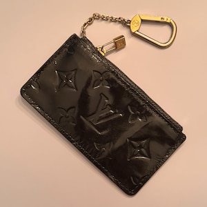 May include: Black patent leather cardholder with embossed floral and logo pattern. Features a gold-tone chain, clasp, and zipper. A luxury accessory for organizing cards and small essentials.