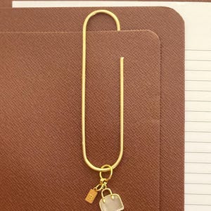 Peut inclure: Un trombone doré avec un petit charme en forme de sac à main et une étiquette avec la lettre "B" attachée. Le trombone maintient une pile de papier ligné blanc et un carnet marron en simili cuir.