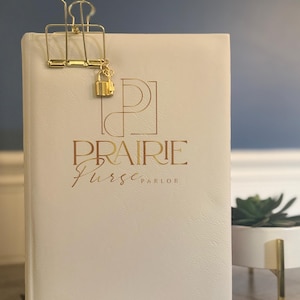 Op de afbeelding: Een crèmekleurig notitieboek met gouden letters met de tekst "PRAIRIE Purse PARLOR". Een gouden klem houdt het notitieboek gesloten, met een kleine gouden hangslotbedel. Het notitieboek ligt op een houten oppervlak.