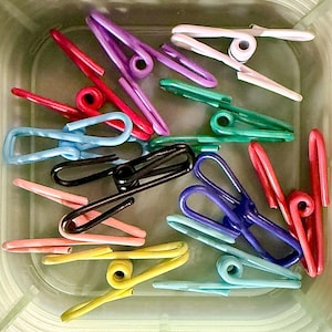 May include: A collection of colorful, plastic clothespins in various shades of red, pink, purple, green, blue, black, and yellow. The clothespins are arranged in a clear, square container, showcasing their diverse hues and designs.