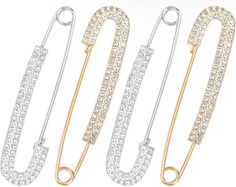 May include: Four large safety pins arranged in a row, alternating between silver-toned metal and gold-toned metal. Each pin features a U-shaped bar encrusted with numerous small, clear, sparkling rhinestones along the entire outer surface.