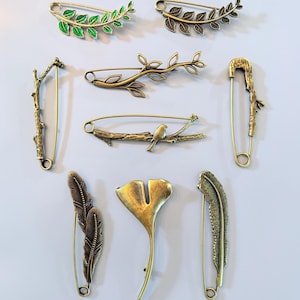 Könnte beinhalten: Eine Sammlung dekorativer Sicherheitsnadeln in verschiedenen von der Natur inspirierten Designs. Die Nadeln weisen Blatt-, Zweig-, Feder- und Vogelmotive in Antikgold- und grünen Emaille-Ausführungen auf. Ideal, um Kleidung oder Accessoires eine einzigartige Note zu verleihen.