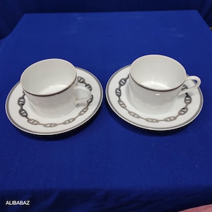 May include: Two white teacups and saucers. Each cup has a dark gray rim and handle. The saucers are white with a dark gray chain link design around the edge. The cups and saucers are on a blue surface.