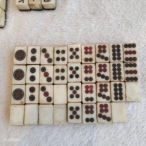 May include: A collection of off-white Mahjong tiles with black and red markings. The tiles are arranged in rows, displaying various symbols and numbers. Some tiles have circular designs, while others feature dots and lines.