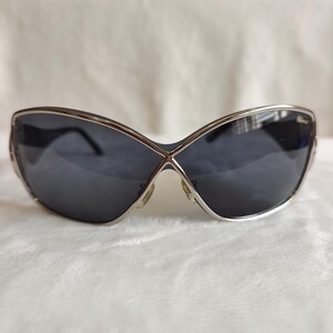 May include: Stylish sunglasses with a silver frame and dark gray lenses. The lenses have a unique shape, with a cross-over design at the bridge. The brand name is visible on the right side of the frame. These sunglasses are a fashionable accessory.