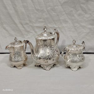 Può includere: Un servizio da tè argentato con intricati motivi floreali. Il set include una teiera, una lattiera e una zuccheriera, tutti con manici e coperchi ornati. La teiera ha un beccuccio curvo e poggia su una base decorativa.