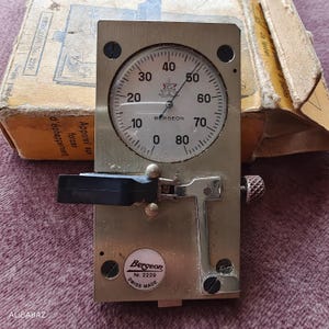May include: A vintage Bergeon watch timing tool, model Nr. 2229, with a circular gauge displaying numbers from 0 to 80. The tool is made of metal and has a black lever and a textured knob. The original box is visible in the background.