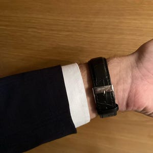 Puede incluir: Un reloj de cuero negro con hebilla plateada abrochado en una mu&ntilde;eca. El reloj se combina con una chaqueta de traje azul marino y un pu&ntilde;o de camisa blanca. La correa del reloj tiene un patr&oacute;n texturizado.